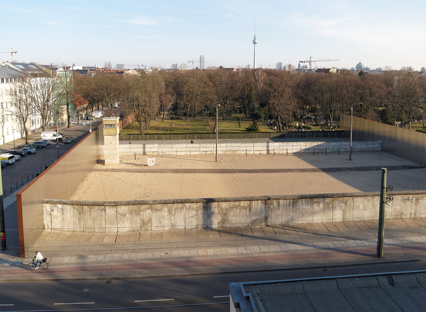 The preserved Berlin Wall border strip at Bernauer Strasse seen from the viewing tower, showing the outer wall, the raked sand death strip, a watchtower and the hinterland wall, with the Berlin TV Tower on the skyline.
